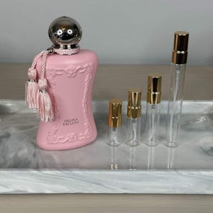 May include: A pink perfume bottle with a silver cap and pink tassels, alongside several clear glass vials with gold caps. The bottle is labeled "DELINA EXCLUSIF" and sits on a marble-patterned tray.