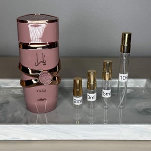 K&ouml;nnte beinhalten: Eine rosa Parf&uuml;mflasche mit goldenen Akzenten und dem Wort "YARA" in Goldbuchstaben. Daneben befinden sich kleine Parf&uuml;mprobenflaschen mit den Aufschriften 2 ml, 3 ml, 5 ml und 10 ml.