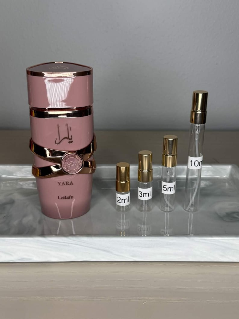 K&ouml;nnte beinhalten: Eine rosa Parf&uuml;mflasche mit goldenen Akzenten und dem Wort "YARA" in Goldbuchstaben. Daneben befinden sich kleine Parf&uuml;mprobenflaschen mit den Aufschriften 2 ml, 3 ml, 5 ml und 10 ml.