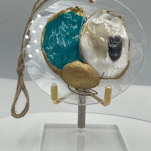 Può includere: Un ornamento circolare trasparente con due conchiglie di ostrica con finiture dorate e una piccola conchiglia dorata. Un guscio è riempito di vernice turchese. L'ornamento è appeso a un anello di spago e una perla dorata. L'ornamento è esposto su un supporto trasparente.