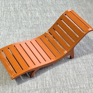 May include: A miniature orange lounge chair featuring a slatted design with a curved backrest and seat. The piece is constructed with a smooth, glossy finish and includes four short legs that elevate the structure from the surface below.