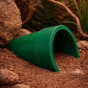 May include: A dark green, hollow, conical-shaped tunnel structure with a wide, circular opening at one end and a tapered, closed end. The object features a smooth, matte surface and is positioned horizontally on a textured, brown, granular surface.