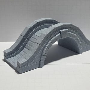 May include: A gray, miniature stone bridge with an arched design. The bridge has a textured surface, with steps leading up to the top. The bridge is a decorative item, suitable for model railroads or tabletop gaming.