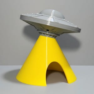 May include: A gray and yellow UFO-shaped object featuring a smooth, conical yellow base with a wide arched opening and a detailed gray saucer top with a central dome and raised rectangular panel accents along the outer rim of the disc structure.