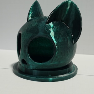 May include: A dark emerald green, faceted cat-shaped object with large pointed ears and a circular opening on the front. The item features a smooth, reflective finish and is mounted on a circular base, displaying a distinct 3D printed texture.