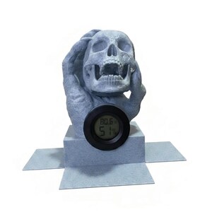 May include: A gray skull sculpture with hands covering its head, holding a digital display. The display shows the temperature at 80.6 degrees and humidity at 51%. The sculpture sits on a gray base.