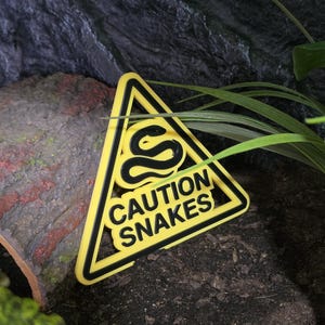 May include: A triangular yellow and black sign featuring a raised graphic of a snake and the text CAUTION SNAKES. The sign has a bold black border and a yellow background, with the snake icon and lettering rendered in a raised black relief style.