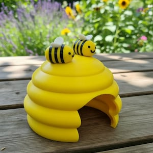 May include: A bright yellow, dome-shaped beehive structure with a smooth, tiered surface and a wide arched opening at the base. Two small, round, yellow and black striped bees with white wings are attached to the top of the hive.