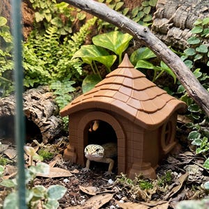 May include: A small leopard gecko with yellow and black spots peeking out from inside a brown, plastic, house-shaped reptile hide. The structure features a shingled roof, a rounded doorway, and a side window, placed on a soil substrate.