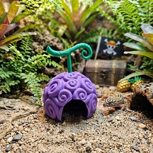 May include: A purple, dome-shaped hideout featuring a textured, swirling pattern and a prominent green, curly stem on top. The object has a hollow arched opening at the base and is positioned on a sandy surface near a small lizard and a pirate flag.