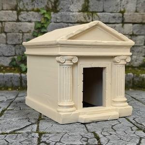 May include: A miniature cream-colored temple structure featuring a triangular pediment roof, two fluted columns with ionic capitals, and a central rectangular doorway opening. The building has a smooth, solid surface with subtle weathered detailing.