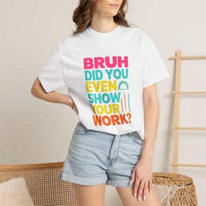以下が含まれることがあります： ピンク、ティール、イエロー、オレンジのカラフルな文字で「BRUH DID YOU EVEN SHOW YOUR WORK?」とプリントされた白い半袖Tシャツ。デザインには、小さなティールの分度器、細いオレンジの定規、ピンクの鉛筆のグラフィックが含まれています。