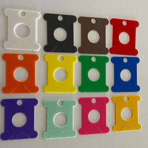 May include: A collection of twelve flat, spool-shaped embroidery floss bobbins arranged in a grid. Each bobbin features a central circular hole, a smaller hole at the top, and two angled slits at the bottom. The colors include white, black, brown, red, orange, yellow, green, blue, purple, light green, pink, and gold.
