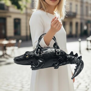 May include: A black handbag shaped like an alligator, with a textured, crocodile-like pattern. The bag has a zipper closure, two handles, and a tail. The alligator's eye is gold. The bag is held by a person wearing a white dress.