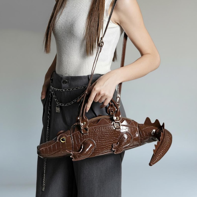 May include: A brown crocodile-shaped handbag with a textured, embossed pattern. The bag has a zipper closure, a top handle, and a long shoulder strap. The bag's design includes a tail, legs, and an eye, giving it a realistic appearance.