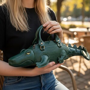 May include: A green alligator-shaped handbag with a textured, crocodile-like pattern. The bag features a top handle, a zipper closure, and gold-colored hardware. The alligator's head and tail are detailed, and the bag is held by a person.