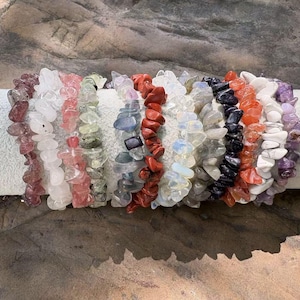 Può includere: Una collezione di braccialetti con schegge di pietre preziose in vari colori, tra cui rosa, bianco, verde, blu, rosso e viola. I braccialetti sono esposti su una superficie cilindrica bianca, mostrando le variazioni naturali delle pietre.