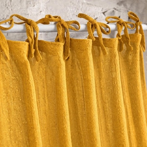 May include: Close-up of mustard yellow curtains. The curtains, made of textured fabric, are tied to a white rod with matching yellow fabric ties. The background is a light-coloured, textured wall.