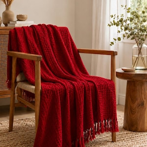 Puede incluir: Una manta de color rojo vibrante con un patrón de tejido de gofre texturizado y flecos anudados a lo largo de los bordes. La manta rectangular está colocada sobre una silla de madera, mostrando su color carmesí profundo y su estructura.
