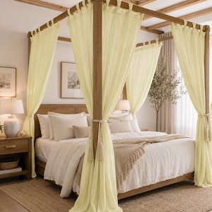 May include: A wooden four-poster bed with cream-colored curtains and bedding. The bed is in a well-lit room with a nightstand, lamp, and framed artwork. The curtains are tied back with tassels, and the bed is covered with a white comforter and a tan throw blanket.