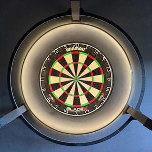 Könnte beinhalten: Ein kreisförmiges Dartboard mit einem schwarz-weißen, segmentierten Design und roten und grünen Akzenten. Das Board wird von einem umlaufenden Licht beleuchtet und ist auf einer dunklen Oberfläche montiert. Der Markenname "Winmau" ist sichtbar.