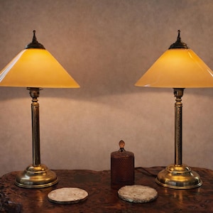 May include: Two matching table lamps with brass bases and stems, topped with amber-colored, triangular shades. A small, dark brown bottle and two round coasters sit on a wooden table. The lamps are lit, casting a warm glow.