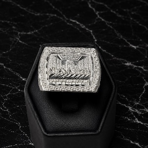 Può includere: Un anello rettangolare color argento con un'incisione in rilievo dettagliata della scena dell'Ultima Cena sulla parte frontale. Il bordo della superficie dell'anello è decorato con un motivo geometrico a greca ripetuto e finitura lucida.