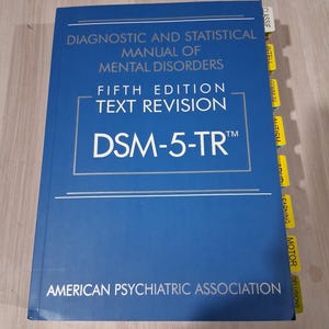Könnte beinhalten: Ein blaues Buch mit dem Titel "Diagnostic and Statistical Manual of Mental Disorders, Fifth Edition, Text Revision, DSM-5-TR" von der American Psychiatric Association. Das Buch hat gelbe Registerkarten und weißen Text.