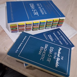 Könnte beinhalten: Eine Sammlung von DSM-5-TR-Büchern, darunter ein großes blaues Lehrbuch mit farbigen Registern und zwei kleinere blaue Nachschlagewerke. Die Bücher liegen auf einer hellen Oberfläche, ideal zum Lernen oder Nachschlagen.