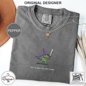 Op de afbeelding: Pepperkleurig t-shirt met een geborduurde cartoonkikker met een tovenaars hoed en cape, met een zwaard. De tekst "OFF TO RUIN THE DM'S PLANS" staat onder de kikker. Het shirt heeft een ronde hals.