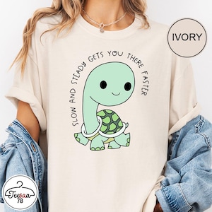Peut inclure: T-shirt ivoire avec un dessin de tortue verte souriante et une carapace à motifs. Le texte "SLOW AND STEADY GETS YOU THERE FASTER" entoure la tortue. Le t-shirt à manches courtes est de couleur crème.