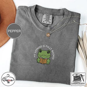 Puede incluir: Camiseta color pimienta con cuello redondo. La camiseta presenta un diseño bordado de una rana verde leyendo un libro, con el texto "MY WEEKEND IS FULLY BOOKED". Un collar plateado con un colgante está sobre la camiseta.