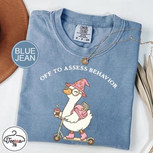 Peut inclure: T-shirt bleu jean avec l'inscription "OFF TO ASSESS BEHAVIOR" et un dessin de canard portant un chapeau de sorcière, des lunettes et des bottes roses, sur un scooter. Le canard tient une tasse de café et un sac à dos. Un collier doré est drapé sur le t-shirt.