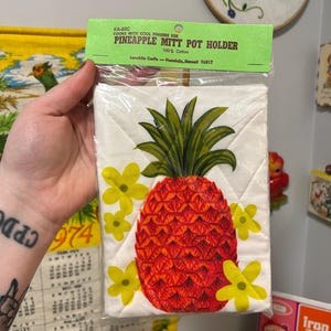 May include: A white quilted pineapple mitt pot holder with a red and yellow pineapple design. The packaging reads "PINEAPPLE MITT POT HOLDER" and "100% Cotton". The pot holder is in a clear plastic package.