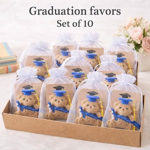 May include: A set of ten graduation favors. Each favor includes a small teddy bear wearing a blue graduation cap and holding a diploma. The bears are packaged in clear bags with white ribbon ties, presented in a brown cardboard tray. The text "Graduation favors Set of 10" is at the top.
