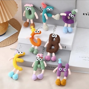 May include: A collection of colorful, crocheted number figures, each with googly eyes, arms, and legs. The numbers are in various colors, including green, orange, yellow, brown, purple, and blue, and are adorned with small hats. These whimsical figures are likely handmade.