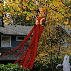 May include: A cylindrical windsock featuring a patterned fabric top with illustrations of black cats, potion bottles, pumpkins, and crescent moons, attached to several long, solid red fabric streamers hanging vertically downward.
