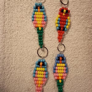 May include: Four handmade keychains crafted from colorful plastic pony beads, each shaped like a small bird. The designs feature various color combinations including blue, yellow, pink, green, red, and orange, with black beads used for the eyes.