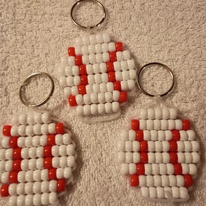 May include: Three circular keychains crafted from white and red plastic pony beads arranged in a grid pattern to resemble baseballs, each featuring a silver metal split ring attached to the top for securing keys or other personal items.