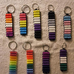 May include: Nine rectangular keychains made of colourful plastic pony beads arranged in vertical stripes. Each keychain features a silver metal split ring at the top, with beads strung on black cord in various colour combinations including rainbow, pride, and flag patterns.