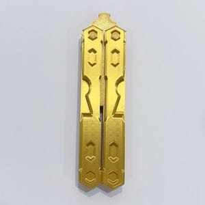 May include: A gold-colored butterfly knife trainer featuring a closed handle design with geometric cutouts and hexagonal screw details. The object has a textured surface finish and a symmetrical structure with two parallel handles joined at the top.
