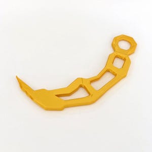 May include: A gold-coloured, curved, rigid object with a pointed tip and a series of geometric cutouts along its length. The handle end features a circular opening, and the overall structure has a layered, textured appearance consistent with 3D printing.