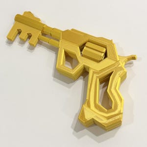 May include: A gold-colored 3D printed object shaped like a revolver with a key-like barrel. The item features a textured, layered surface finish typical of additive manufacturing, with a distinct handle, cylinder, and a multi-pronged key head.