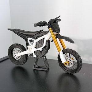 May include: A miniature dirt bike model featuring a white frame, black seat, and black handlebars. The front suspension forks are gold, and the bike is supported by a black stand. It has black knobby tires, silver disc brakes, and a black motor housing.
