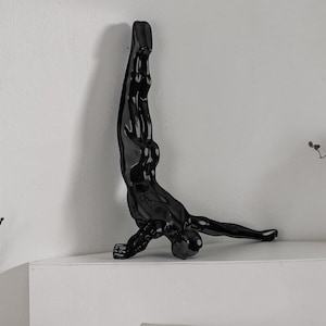 Könnte beinhalten: Eine glänzend schwarze Skulptur einer Person im Handstand. Die Figur ist auf einer weißen Oberfläche montiert, mit ausgestreckten Armen und Beinen. Die Skulptur ist ein dekorativer Artikel für ein Zuhause.