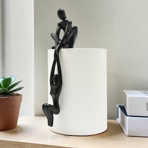 Op de afbeelding: Een witte keramische vaas met twee zwarte abstracte menselijke figuren. Eén figuur zit bovenop de vaas, terwijl de andere aan de zijkant hangt. Links staat een kleine vetplant in een pot.