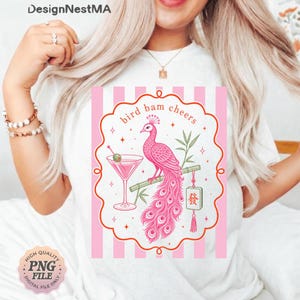 May include: White t-shirt featuring a pink and white graphic. The graphic includes a pink peacock perched on a bamboo branch next to a martini glass. The text "bird bam cheers" is above the peacock. A "High Quality PNG File" logo is in the bottom left.