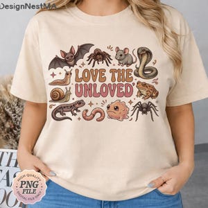 Puede incluir: Camiseta beige con un gráfico colorido de varios animales, incluyendo un murciélago, una araña, un ratón y una serpiente, con el texto "LOVE THE UNLOVED". El diseño tiene un estilo vintage de dibujos animados.