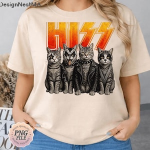 Pode incluir: Camiseta bege com um desenho de quatro gatos ao estilo da banda de rock KISS, com o logotipo da banda "HISS" em laranja e amarelo. Os gatos usam roupas de rock, com um deles mostrando a língua.