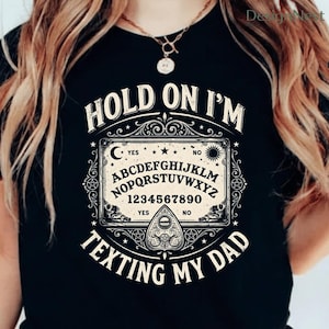 May include: Black t-shirt featuring a white Ouija board design with the text "HOLD ON I'M TEXTING MY DAD". The graphic includes a planchette, moon, sun, stars, and the alphabet.
